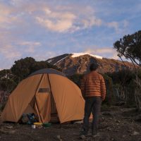 A tent and a hiker in orange admiring the view of Mt. Kilimanjaro.